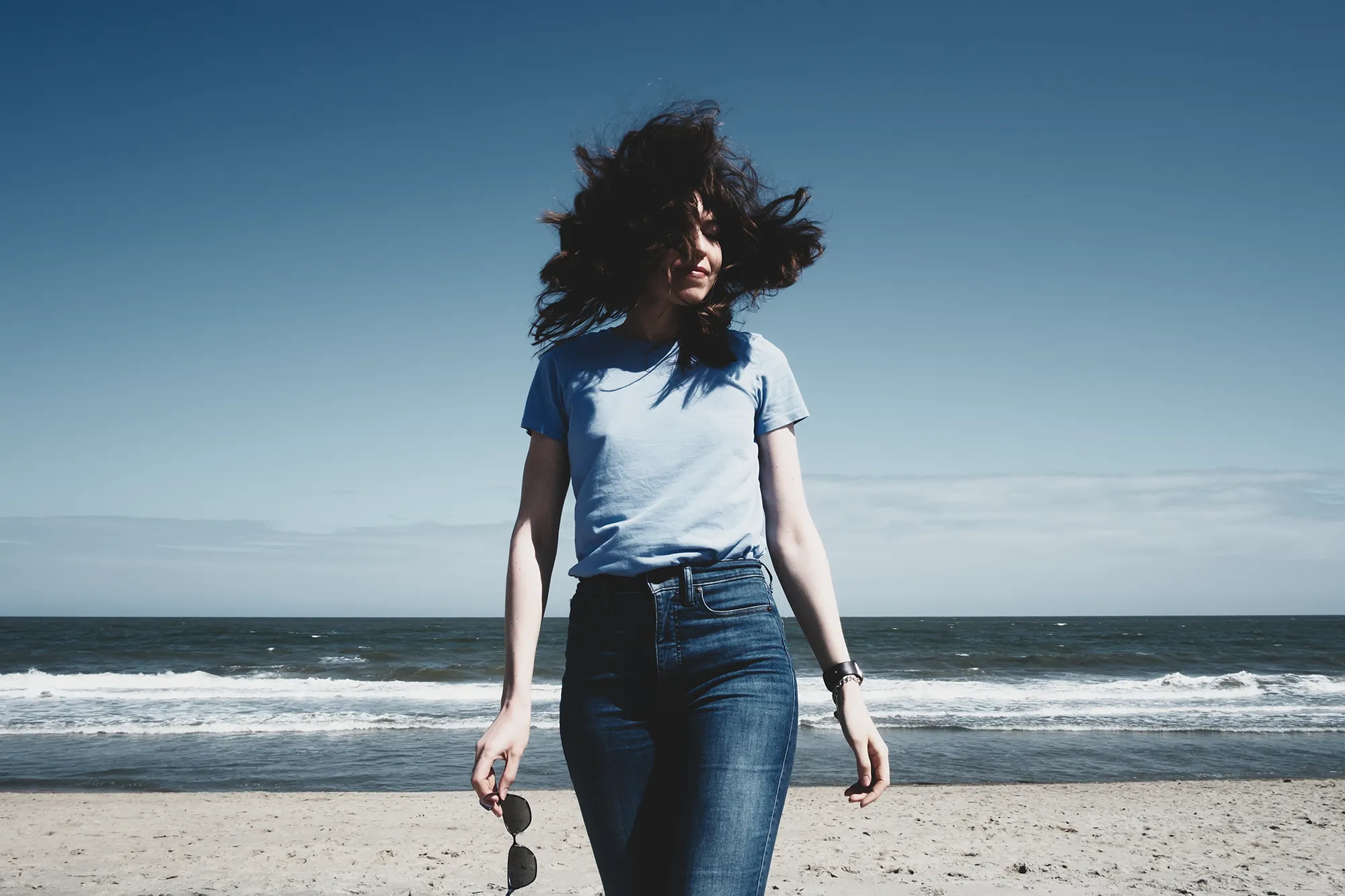 Photo of Oksana at the beach. She's wearing a blue t-shirt and jeans. Her dark brown hair is flying in the wind all over the place, partially covering her face. The waves in the background are calm.